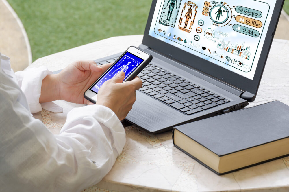 PC am Tisch mit einem Buch davor ein Mensch der ein Handy in der Hand hat