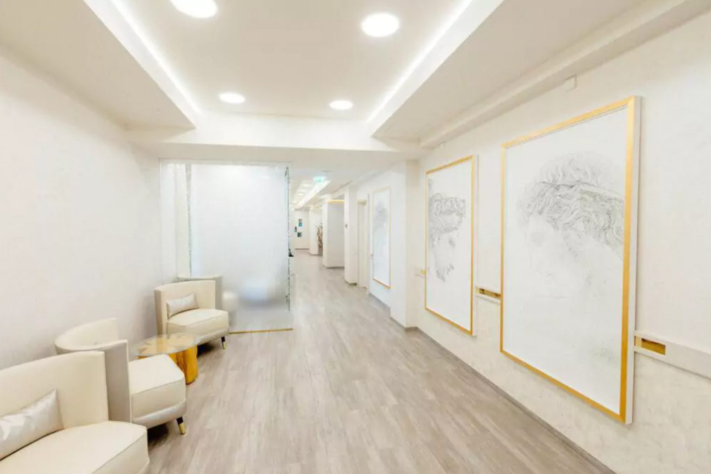 Ästhetische Chirurgie Wien Gang mit hellem Fußboden, weißen Wänden mit großen hellen Bildern an, eine Glasschiebetüre sowie Sitzmöbel in beige