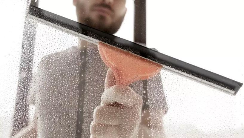 Glasscheibe mit einem großen Fensterwischer und orangem Haltergriff zu sehen der von einem Mann gehalten wird bei dem nur der Oberkörper und der Mund sowie die Nase zu sehen sind