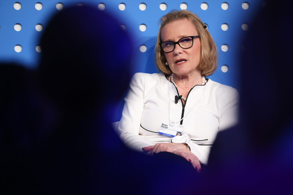 CEO von Merck Belén Garijo in weißer Bluse vor einem blauen Hintergrund in dem weiße Löcher zu sehen sind. 