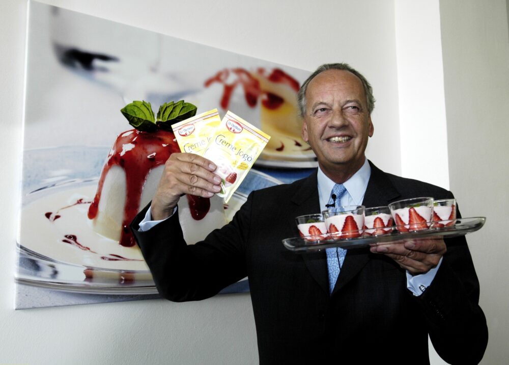 Dr. August Oetker hält Panna Cotta Gläschen und 2 Packungen einer Pulvermischung in der Hand und lächelt