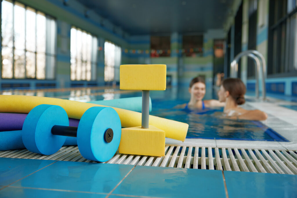 Zwei Frauen befinden sich in einem Schwimmbad. Am Schwimmbadrand liegen Hanteln und Schwimmutensilien.