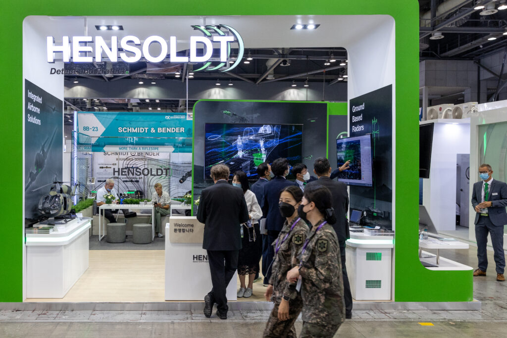Messestand der Firma HENSOLDT mit grünem Rand um den Eingang und viele Menschen mit Anzügen und Mundschutzmasken