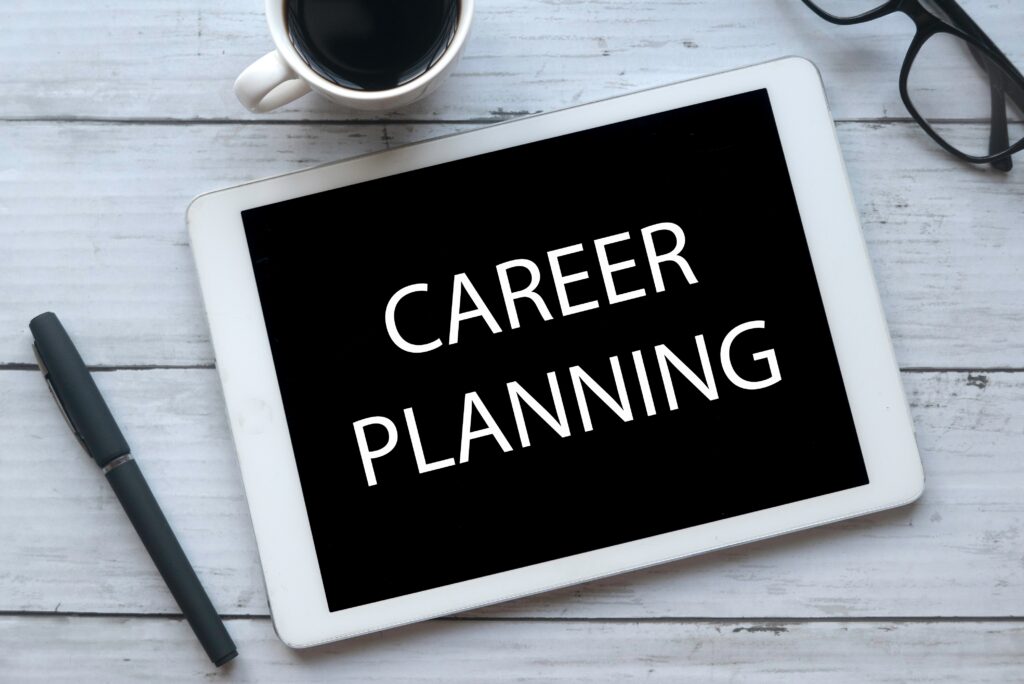 Bernard KRONE Hintergrund von einem hellen Holztisch mit einer Kaffeetasse, einer Brille, einem schwarzen Stift und einem Tablet mit schwarzem Hintergrund auf dem Career Planning steht