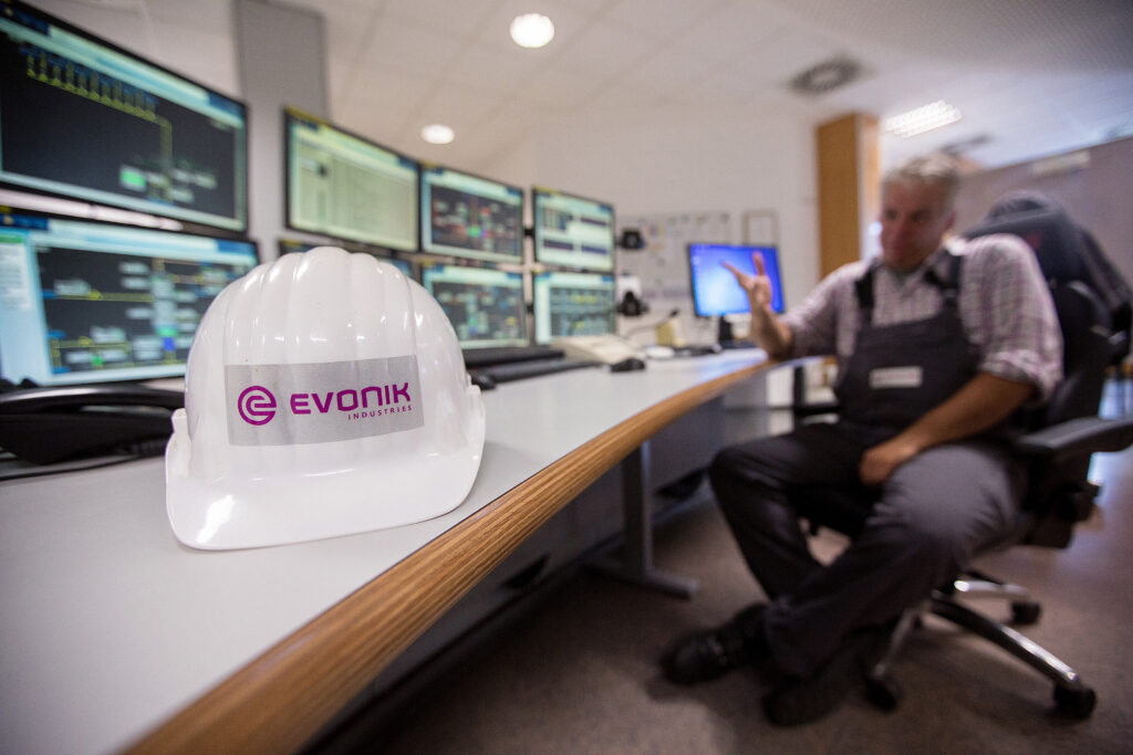 Arbeitshelm von Evonik liegt auf einem Tisch. Dahinter sitzt ein Mitarbeiter in Arbeitsmontur