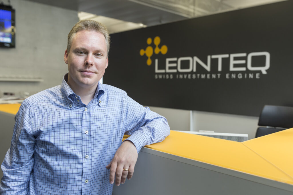 Mann mit blau kariertem Hemd lehnt an einem Stehpult. Dahinter das Logo von Leonteq
