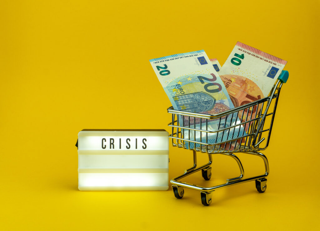 gelber Hintergrund mit dem beleuchteten Schild "Crisis" und einem kleinen Einkaufswagen mit Euro-Scheinen