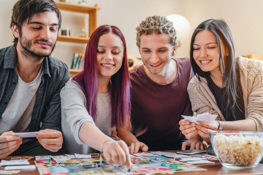 Vier junge Menschen sitzen an einem Tisch und spielen gemeinsam. Das Brettspiel liegt auf dem Tisch. Daneben eine Glasschale mit Popcorn